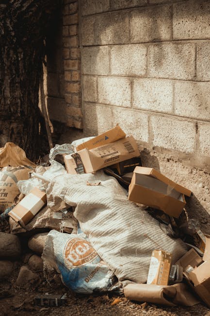 A pile of mixed waste consisting of black plastic garbage bags, a yellow plastic container, and a discarded old car tire are shown in a gravel driveway area. The waste is surrounded by a low stone wal